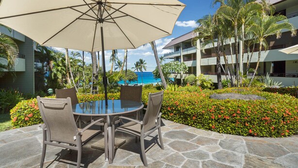 A table with four chairs and a large umbrella in an outdoor setting with a view of a building and a pool. - Summer Deal White Sands Beach Condo with Pool Lanai and Ocean Views (Kailua Kona)