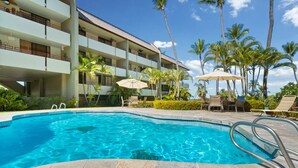 A swimming pool with a stone border, surrounded by lounge chairs and umbrellas, with a multi-story building in the background. - Summer Deal White Sands Beach Condo with Pool Lanai and Ocean Views (Kailua Kona)