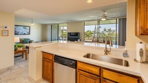 A modern kitchen with a dishwasher, a sink, and a large island. There is a dining area with a table and chairs, and a television in the background. - Summer Deal White Sands Beach Condo with Pool Lanai and Ocean Views (Kailua Kona)
