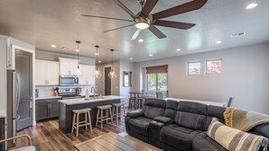 A modern kitchen with a central island, wooden stools, a dark leather sofa, and a ceiling fan. - Steps to Pool - Modern 3BR Golfside w/ Fire Pit (St. George)