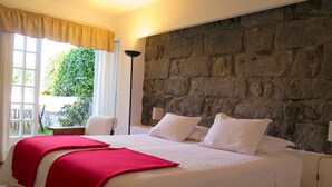A bedroom with a stone wall, a bed with white bedding and red accents, a chair, and a window with curtains. - Quinta das Buganvílias (Horta)