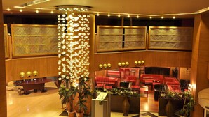 Um saguão de hotel com balcão de recepção, área de estar, luminárias decorativas e uma grande planta.