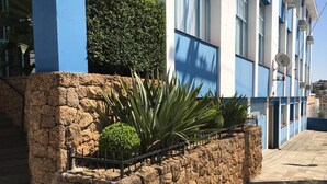 Una jardinera de piedra con plantas y una barandilla de metal, junto a un edificio con paredes azules y blancas y un cielo despejado.