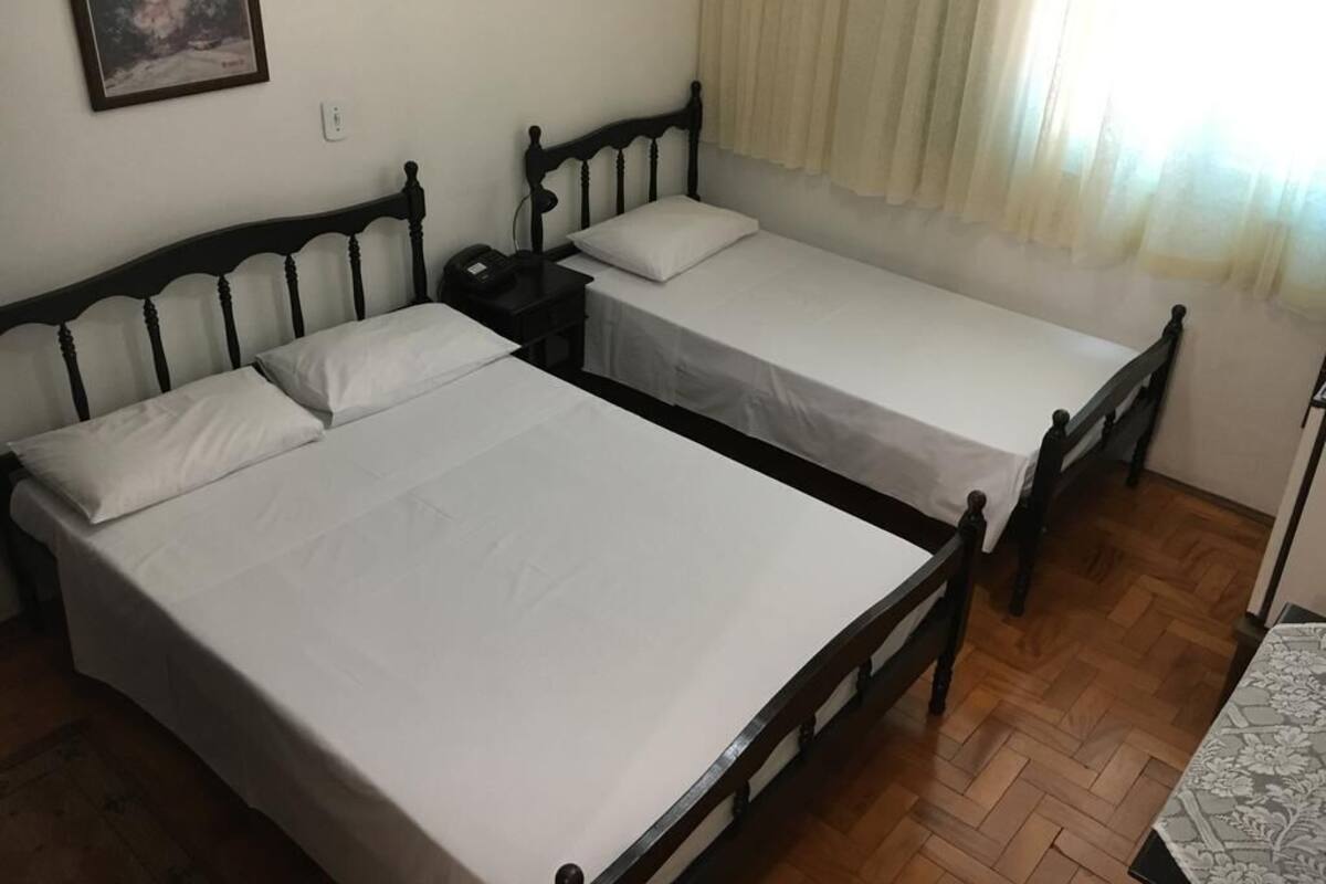 Quarto com duas camas de solteiro, mesa de cabeceira, luminária e janela com cortinas.