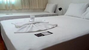 Habitación de hotel con dos camas, ropa de cama blanca, una mesita de noche con lámpara y una pequeña tarjeta sobre la cama.