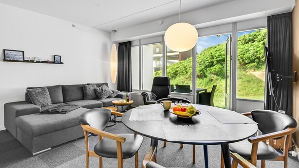 A modern living room with a grey sofa, a round dining table with chairs, a large window with a view of greenery, and a pendant light. - Awesome apartment in Ringkøbing (Ringkøbing)