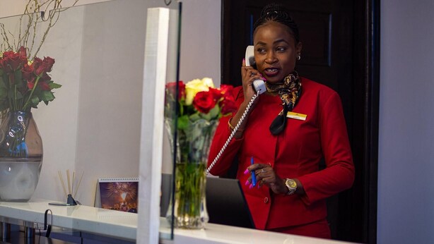 A woman in a red suit standing at a reception desk with a phone, a vase of flowers, and a framed picture on the counter. - The Nest Boutique Hotel (Naivasha)