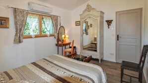 Un dormitorio con cama, una silla, tocador con espejo, una puerta y un equipo de aire acondicionado.
