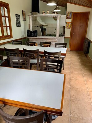A restaurant with a long counter, multiple tables, and chairs. There is a kitchen area with a stove and overhead lighting.