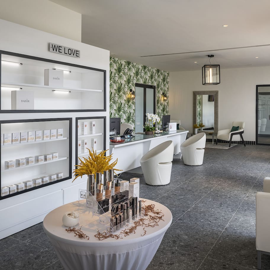 A modern hotel lobby with a reception desk, white sofas, and a display of products on shelves.