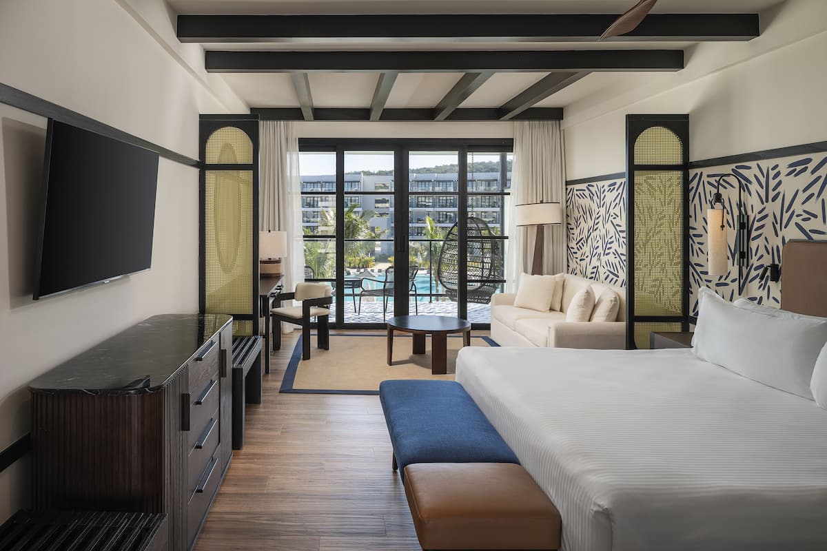 A hotel room with a large bed, a TV mounted on the wall, a seating area with a table and chairs, and a view of the pool and buildings outside.