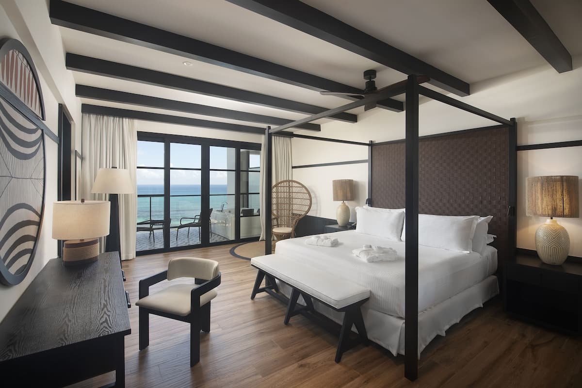 A modern hotel room with a large bed, a desk, a chair, and a balcony with a view of the sea.