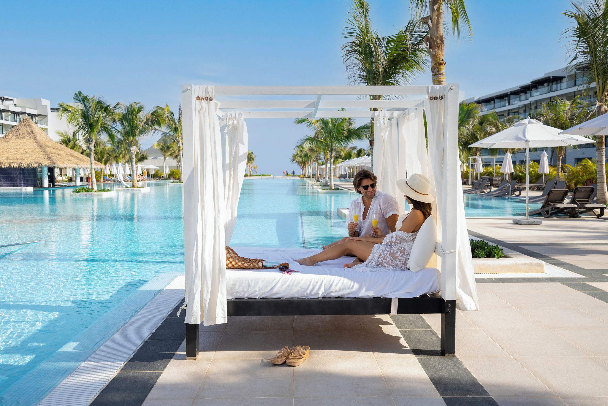 A couple relaxing on a white canopy bed by a pool with palm trees and a clear sky in the background.