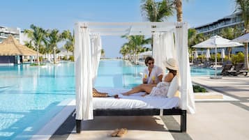A couple relaxing on a white canopy bed by a pool with palm trees and a clear sky in the background.