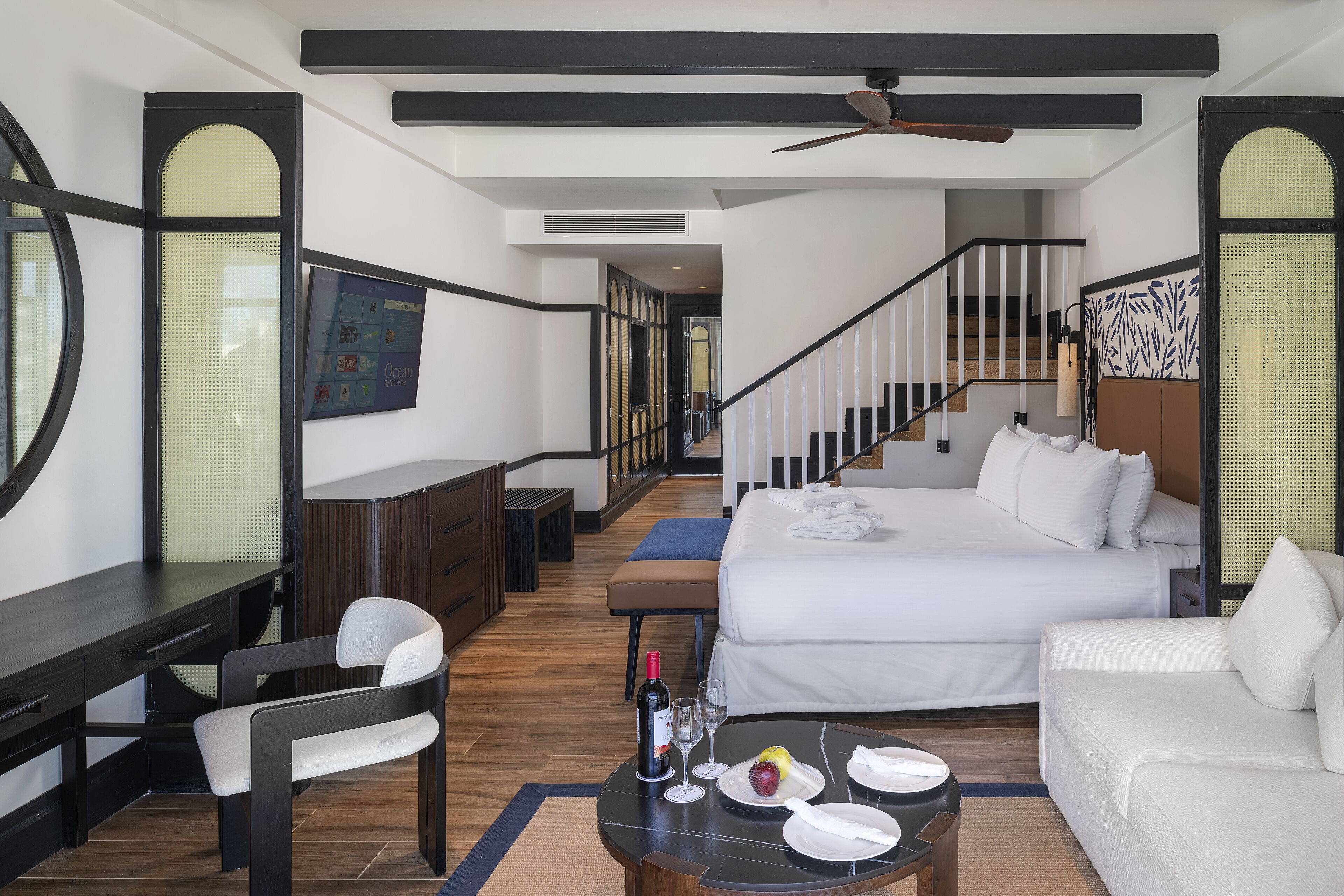 Una camera d'albergo con un letto, una scrivania, una sedia, un tavolino da caffè con una bottiglia e dei bicchieri, e una televisione appesa al muro.
