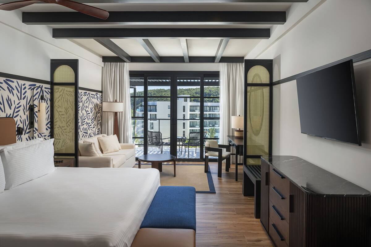 A hotel room with a large bed, a desk, a TV, and a view of a pool and buildings.