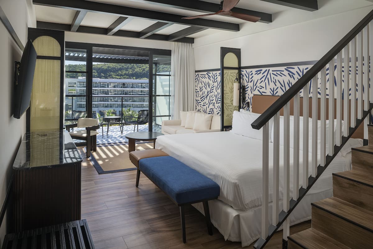 A modern hotel room with a large bed, a bench, a desk, and a balcony with a view of buildings and greenery.