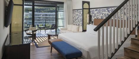 A modern hotel room with a large bed, a bench, a desk, and a balcony with a view of buildings and greenery.