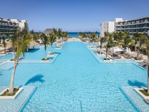 A resort pool with clear blue water, surrounded by palm trees and lounge chairs, with a view of the ocean and modern buildings in the background. - Ocean Eden Bay - Adults Only - All inclusive (Falmouth)