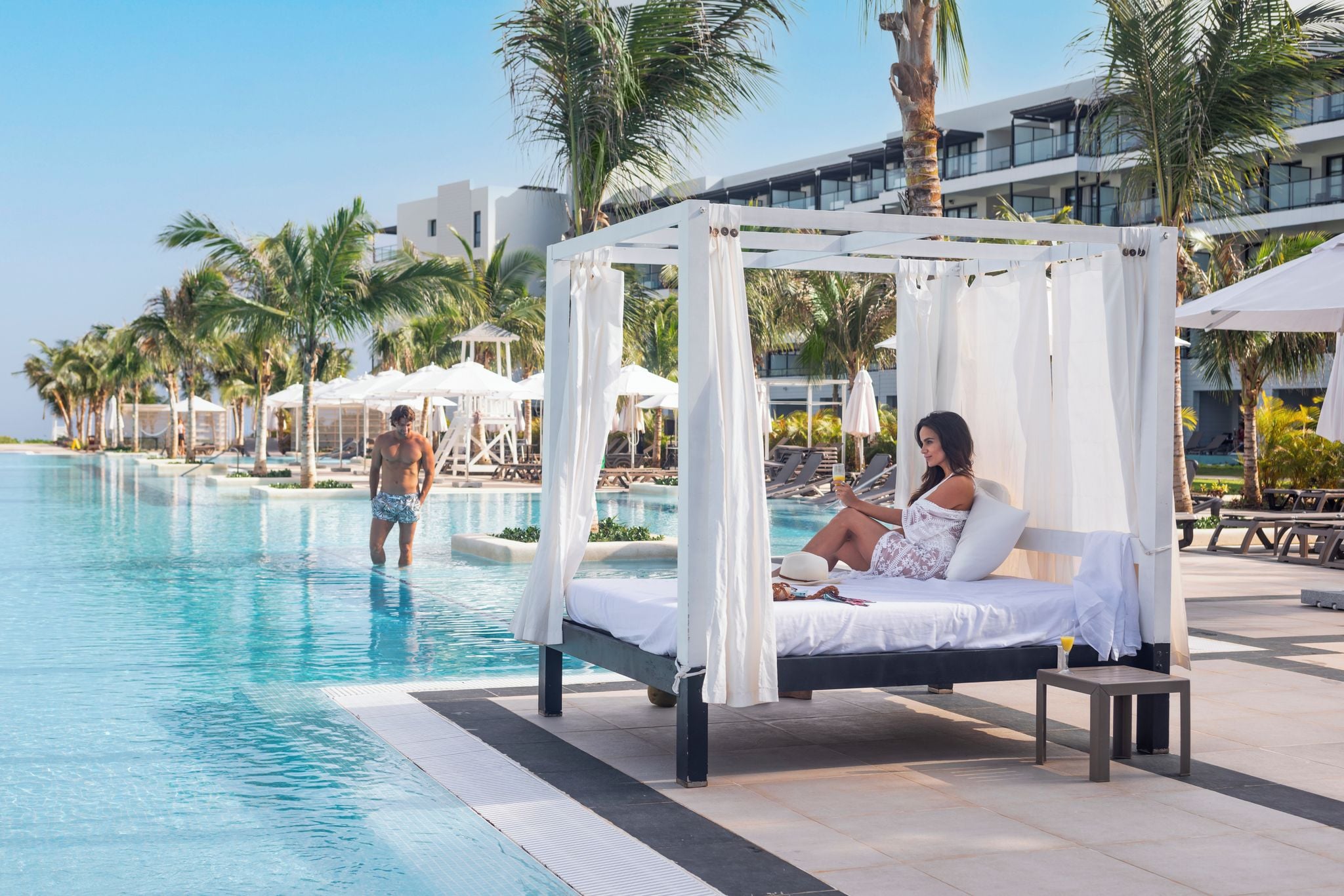 A poolside bed with a canopy, a lounge area with umbrellas, and a pool with palm trees in the background.