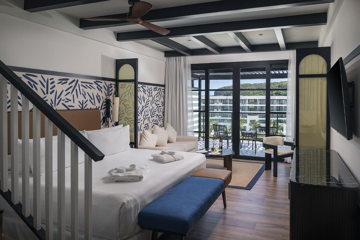 A modern hotel room with a large bed, a sofa, a desk, and a balcony with a view of the city.
