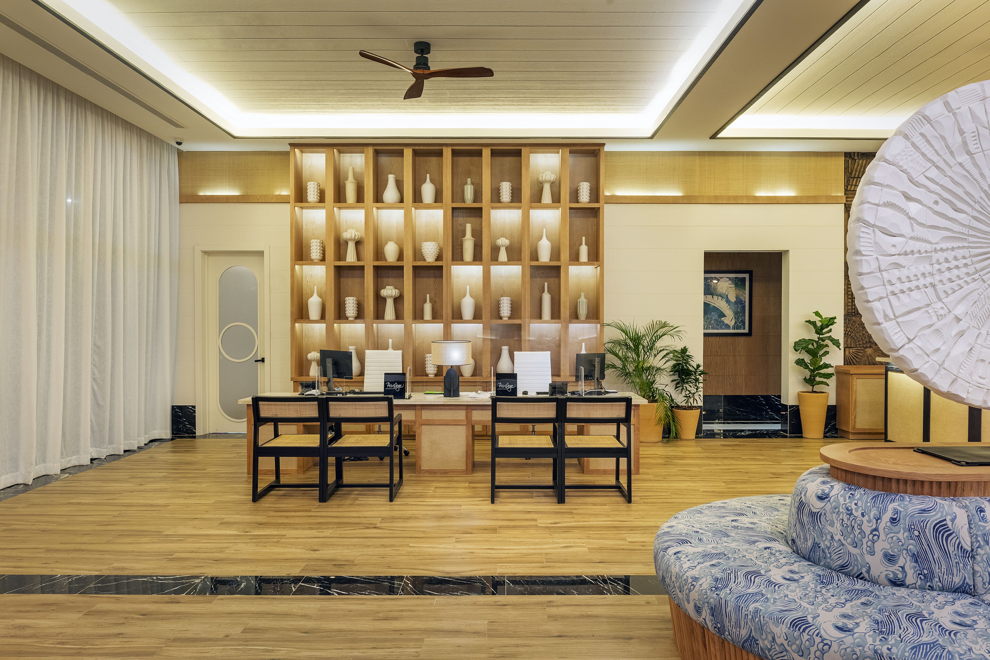 A reception area with a wooden desk, two chairs, a large decorative fan, and a seating area with a patterned sofa.