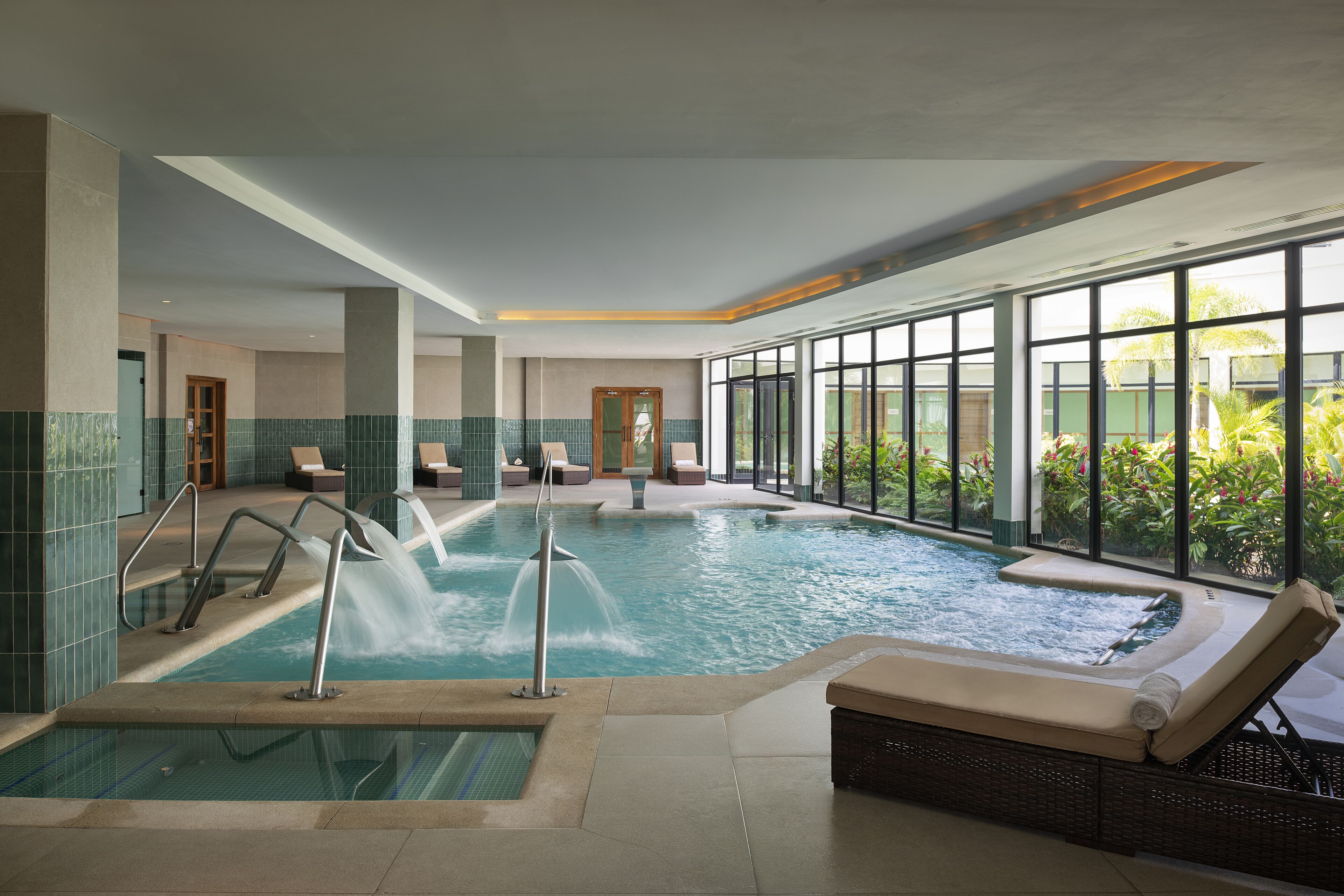 A large indoor swimming pool with a jacuzzi area, lounge chairs, and a view of greenery outside.