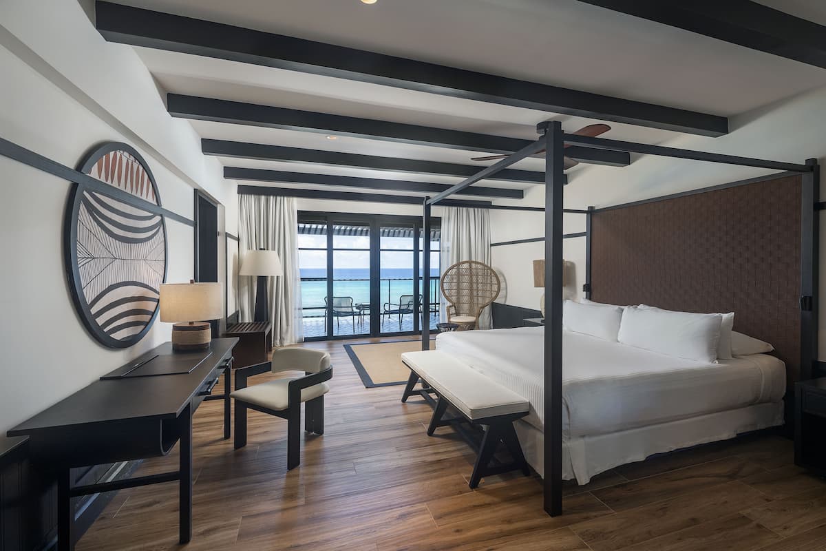 A modern hotel room with a large bed, a desk, a chair, and a view of the sea through large windows.