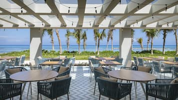 An outdoor seating area with tables and chairs, overlooking a beach with palm trees and the ocean.