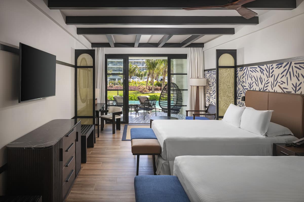 A hotel room with a large bed, a desk, and a TV. There is a balcony with outdoor seating and a view of the hotel and greenery.