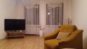 A living room with a flat-screen TV on a wooden stand, a mustard-colored armchair, a white vase with dried branches, sheer curtains, and a table lamp.