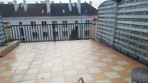 A tiled balcony with a metal railing, a wicker chair, and a view of a building with pink trim and multiple windows.