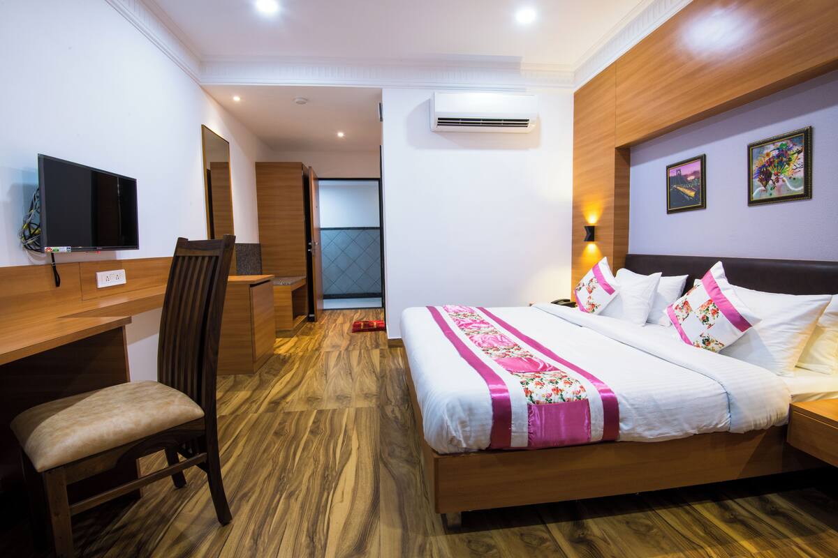 A hotel room with a wooden desk, a chair, a bed with white bedding and pink accents, a flat-screen TV mounted on the wall, and an air conditioning unit.