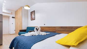 A modern hotel room with a large bed, a wooden headboard, a blue bedspread, a yellow pillow, and a heart-shaped towel on the bed.