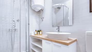 Un bany modern amb una dutxa de vitrall, un lavabo blanc amb taulell de fusta i un mirall sobre el lavabo.