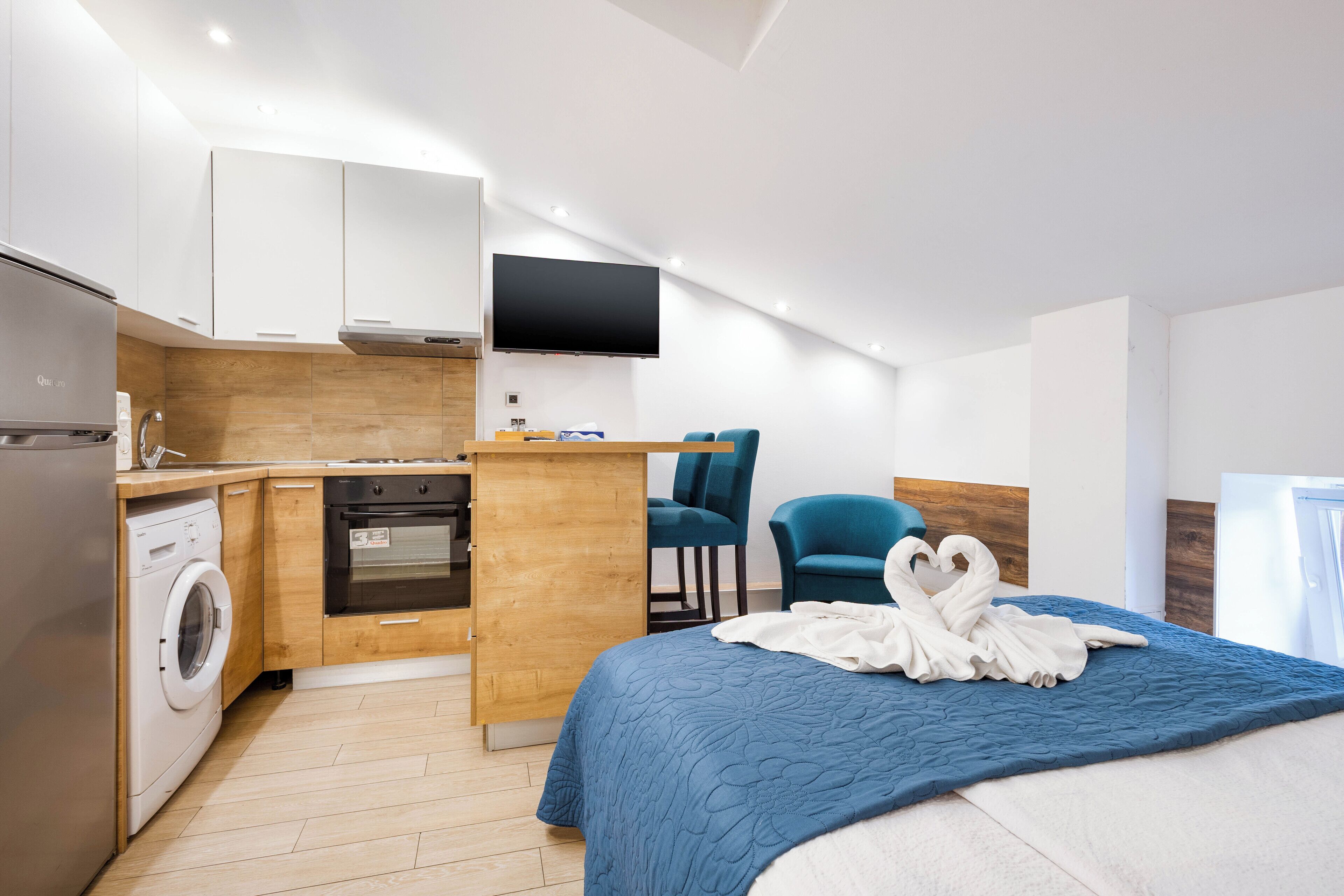 A modern hotel room with a kitchen area, a bed with a blue quilt, and a flat-screen TV mounted on the wall.
