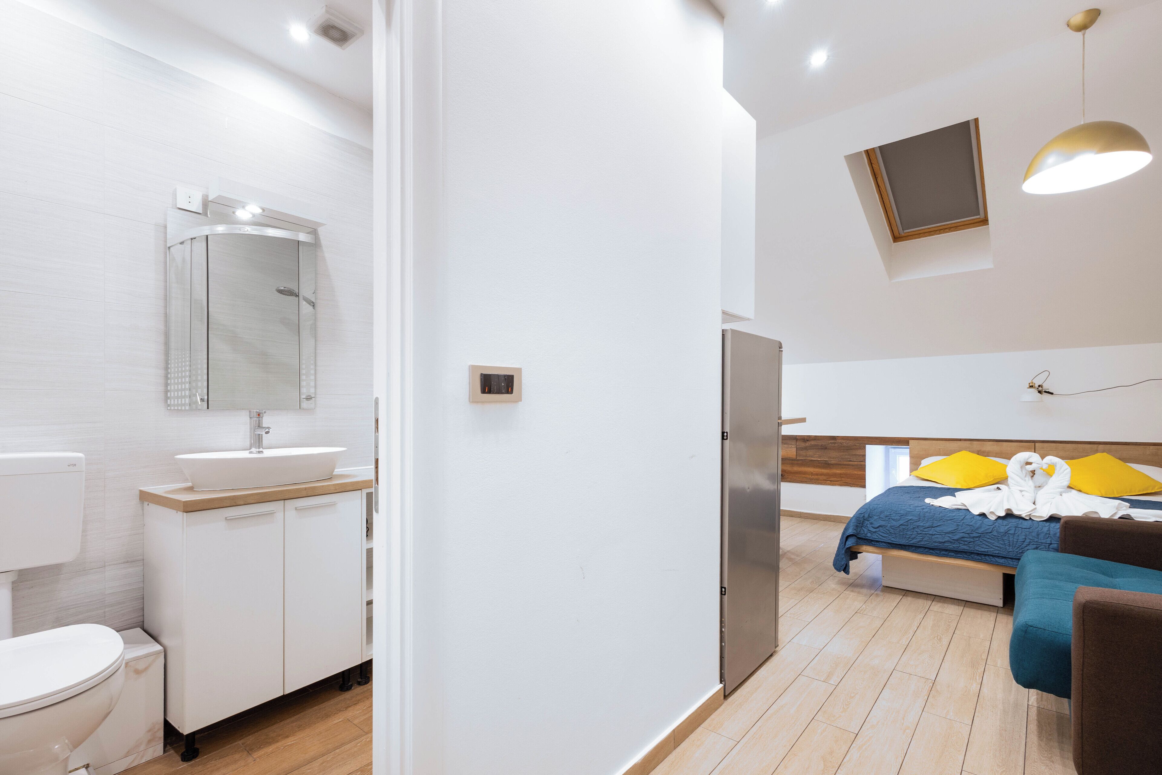 A modern hotel room with a bathroom, a bed with yellow pillows, and a TV mounted on the wall.