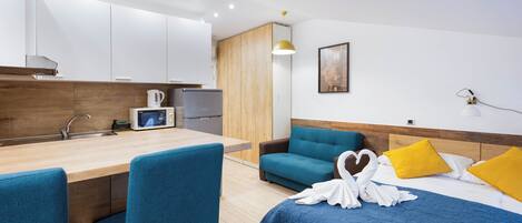 A modern hotel room with a kitchenette, a bed with blue and yellow pillows, and a blue chair.