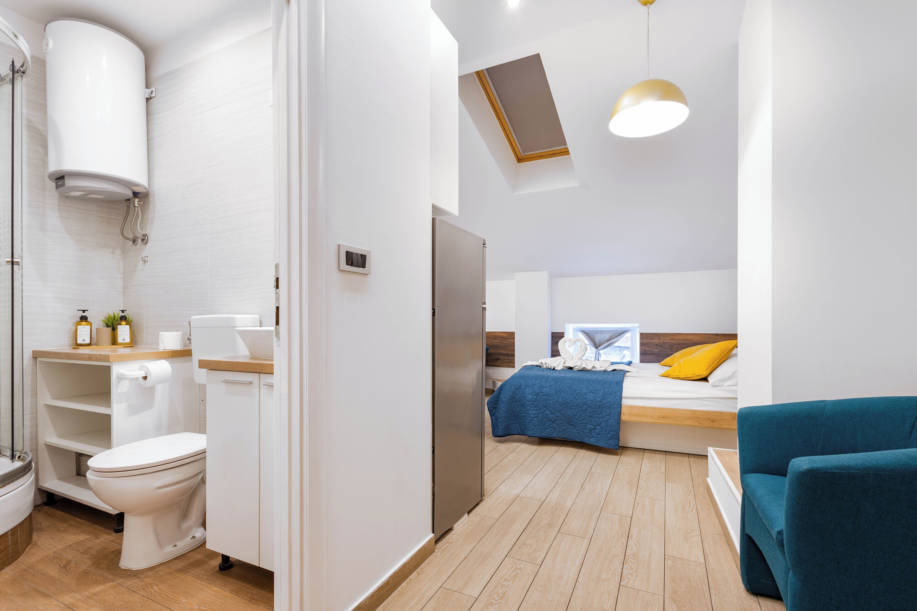 Ein modernes Badezimmer mit Toilette, Waschbecken und Dusche. Ein Bett mit blauer Decke und gelbem Kopfkissen ist ebenfalls zu sehen. Ein türkisfarbenes Sessel steht im Raum.