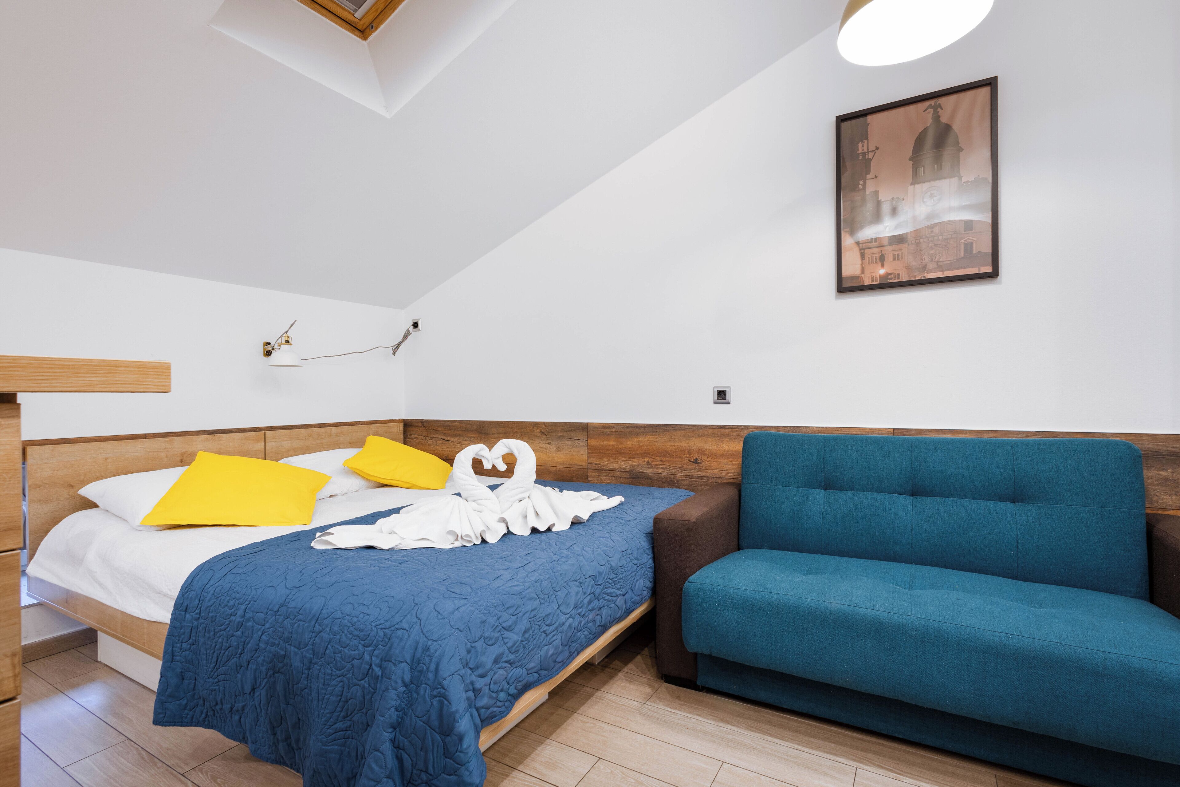A bedroom with a bed, a blue sofa, a wooden headboard, a white wall, a framed picture, and a ceiling light.