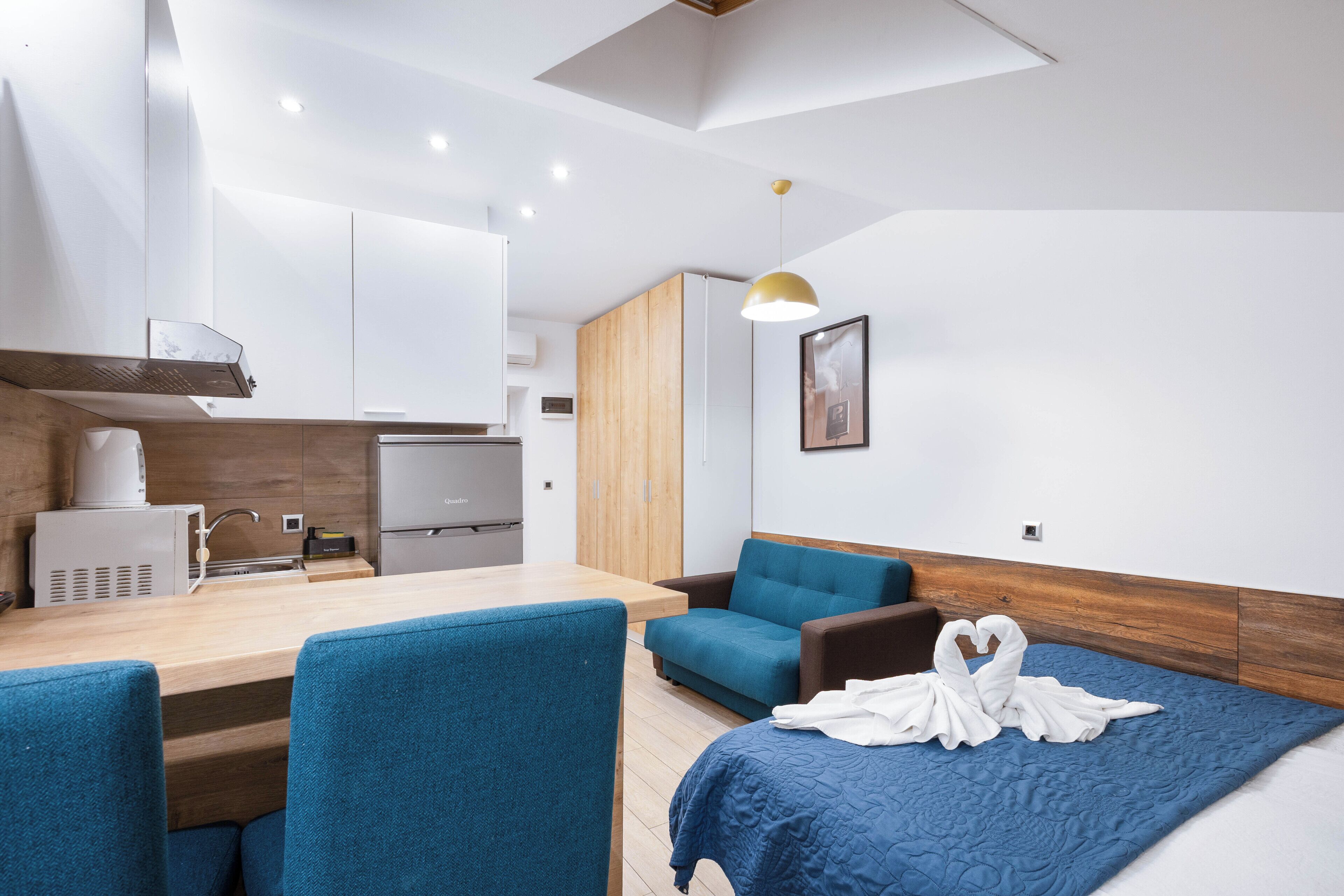 Une chambre d’hôtel moderne avec une kitchenette, un lit recouvert d’une couette bleue et un canapé bleu.