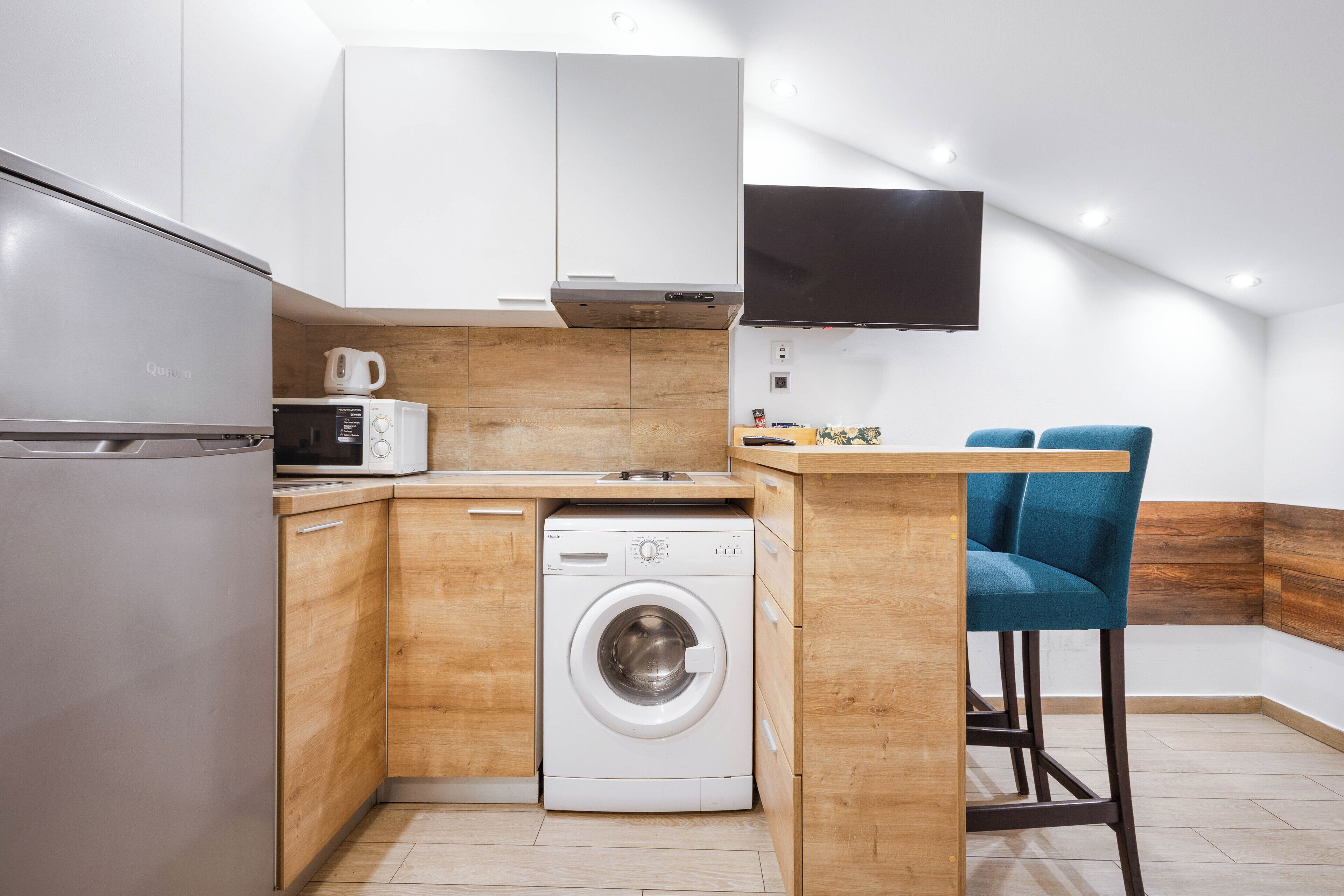 A compact kitchen with a washing machine, microwave, and a flat-screen TV mounted on the wall.