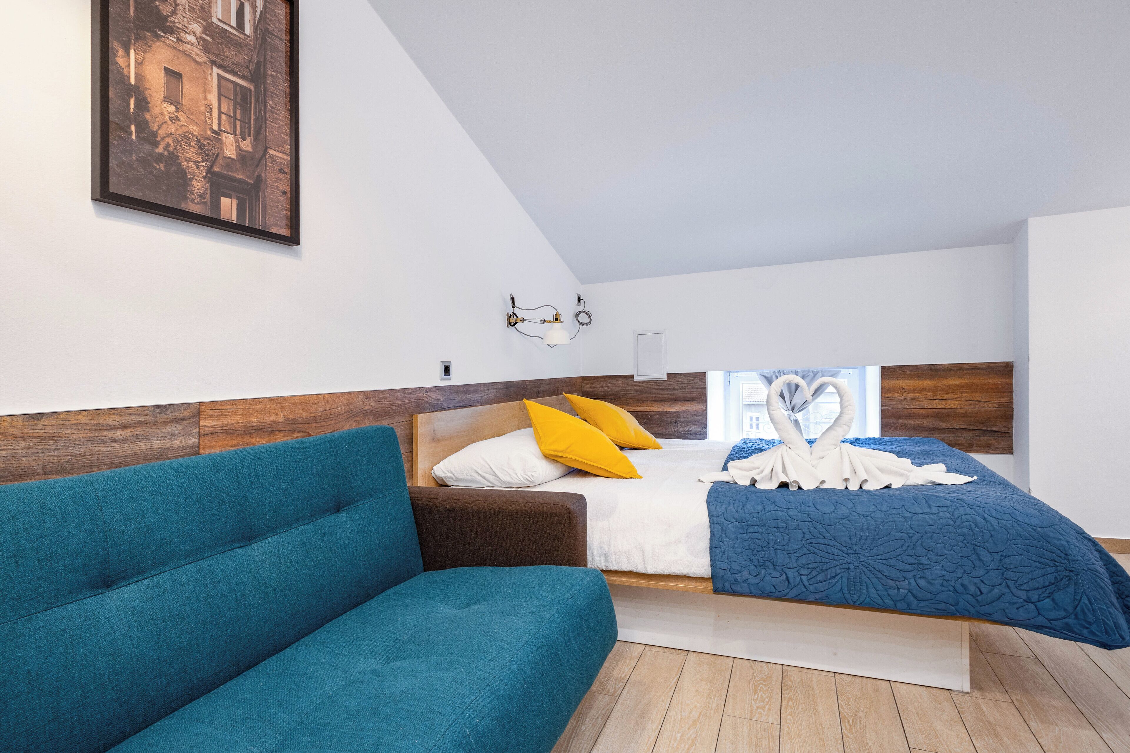 A modern bedroom with a wooden headboard, a bed with a blue quilt, a teal sofa, and a framed picture on the wall.