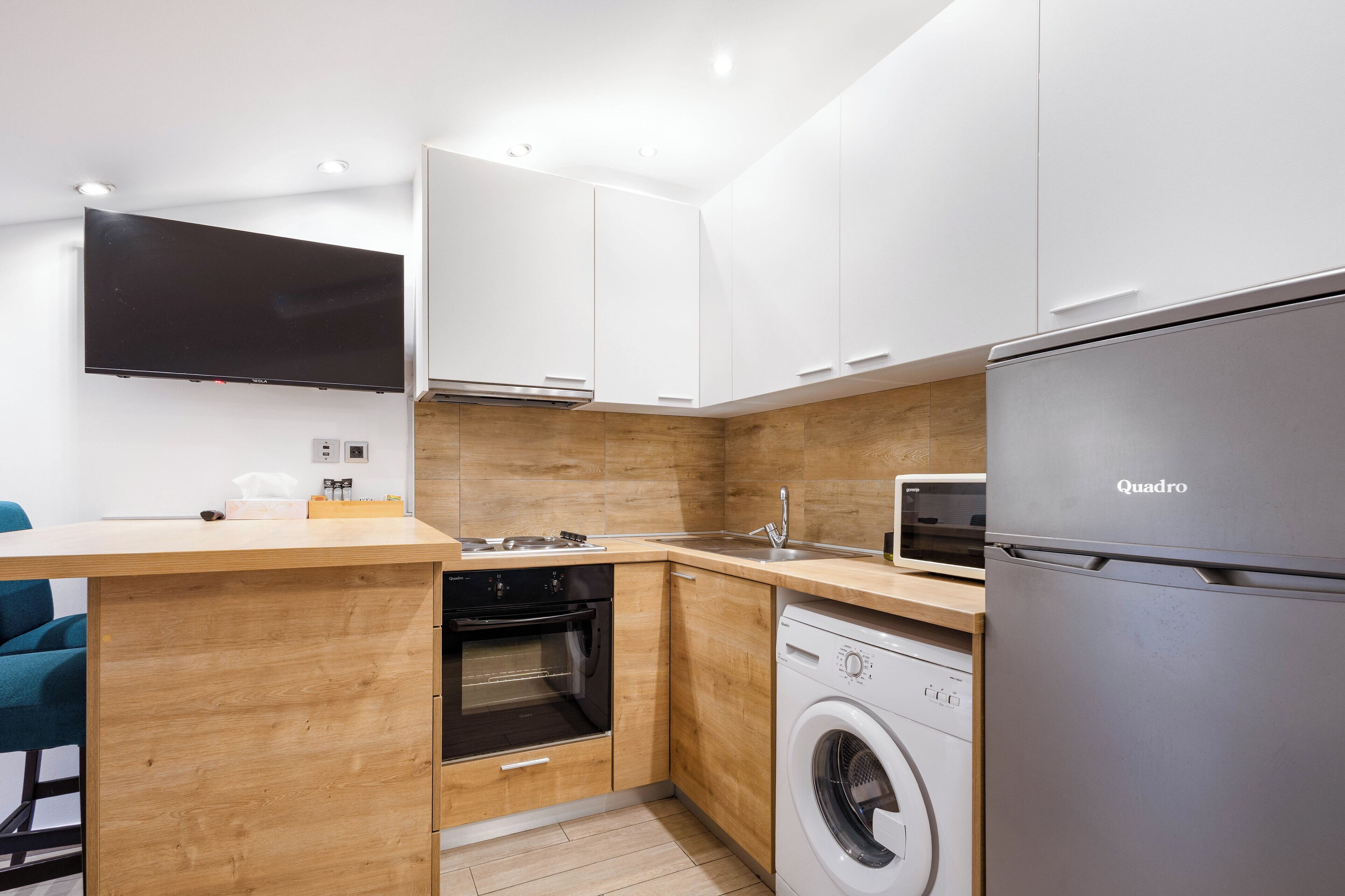 A compact kitchen with a built-in oven, microwave, washing machine, and a large flat-screen TV.