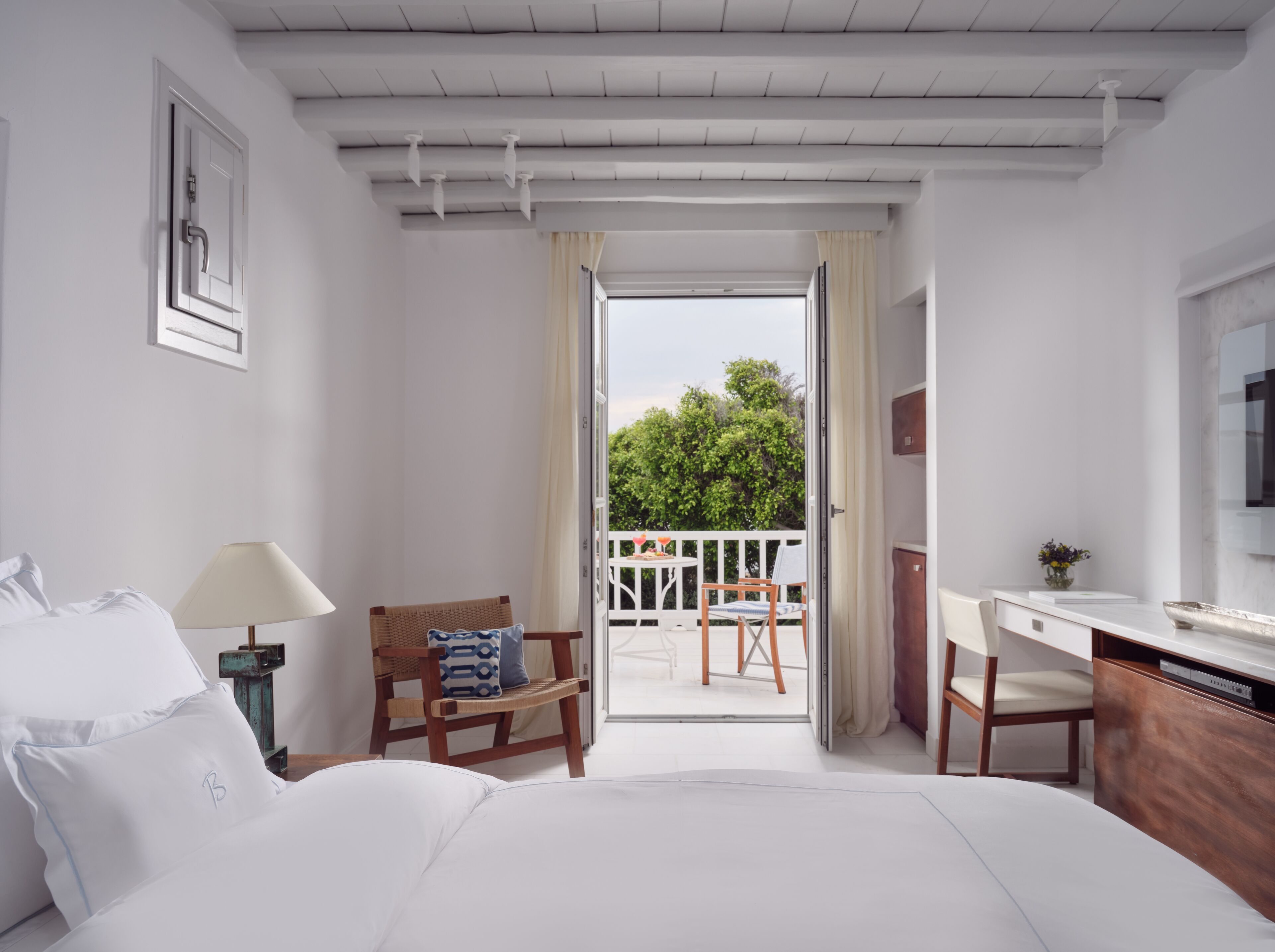 A bedroom with a bed, a desk, a chair, and a balcony with a view of greenery.