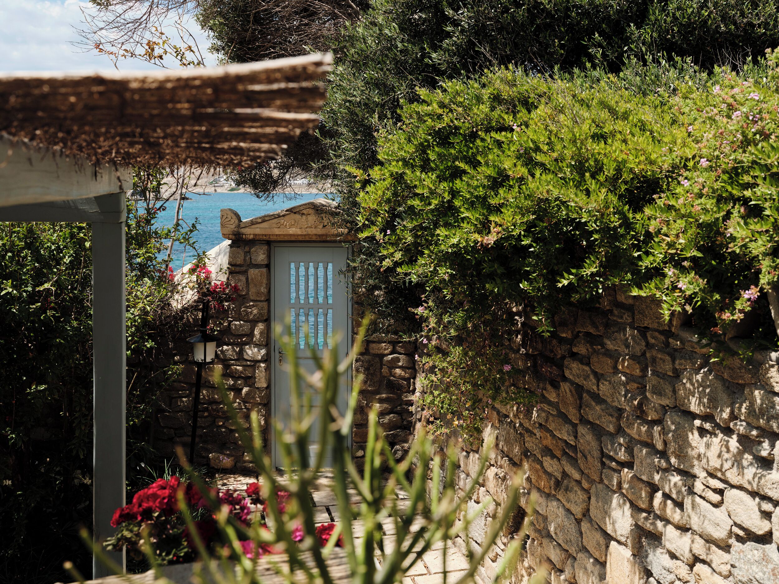 A stone wall with a door, lush greenery, and a view of the sea.
