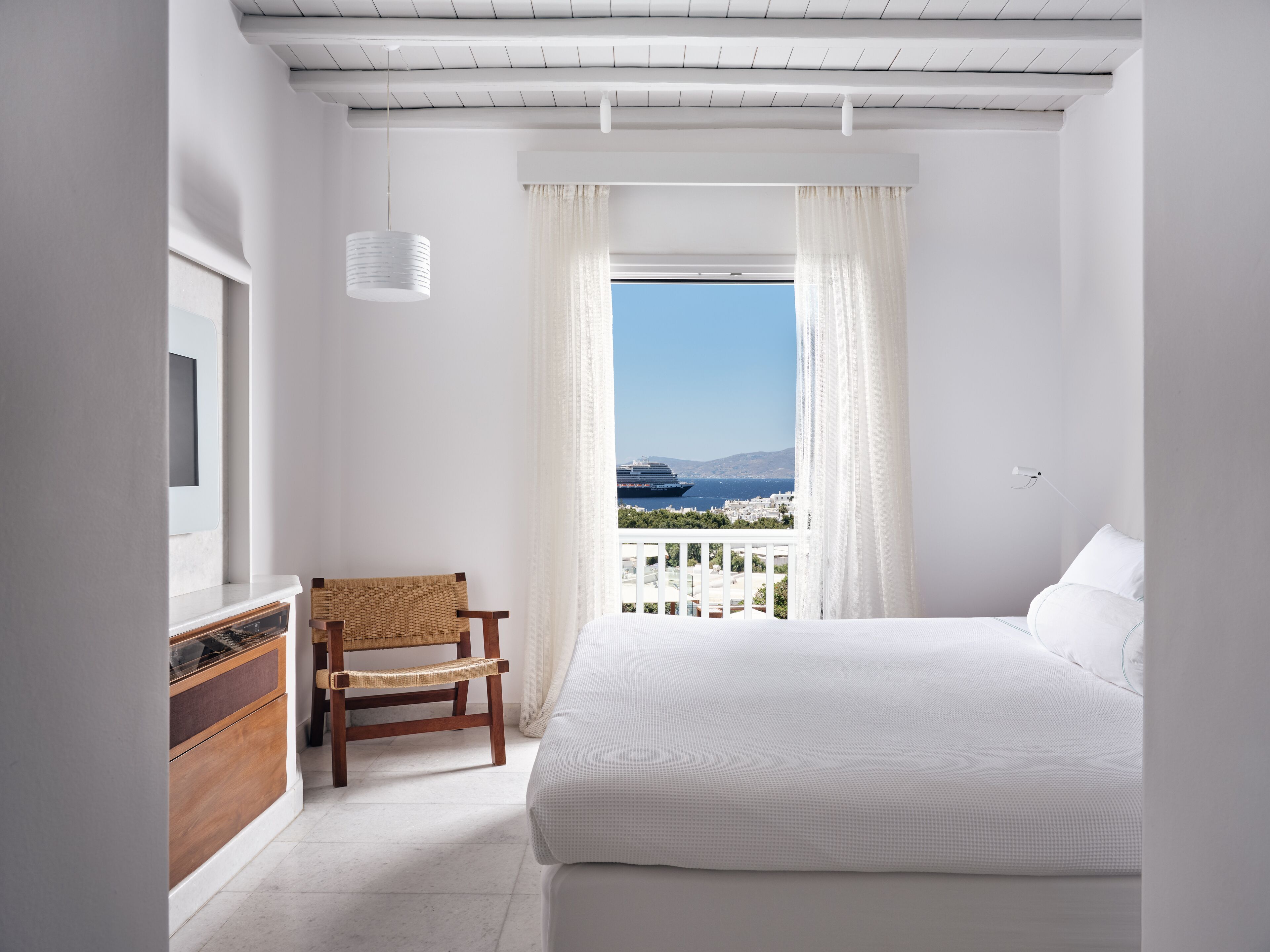Een slaapkamer met een groot bed, een houten stoel, een dressoir en een raam met uitzicht op een schip en stadsgezicht.