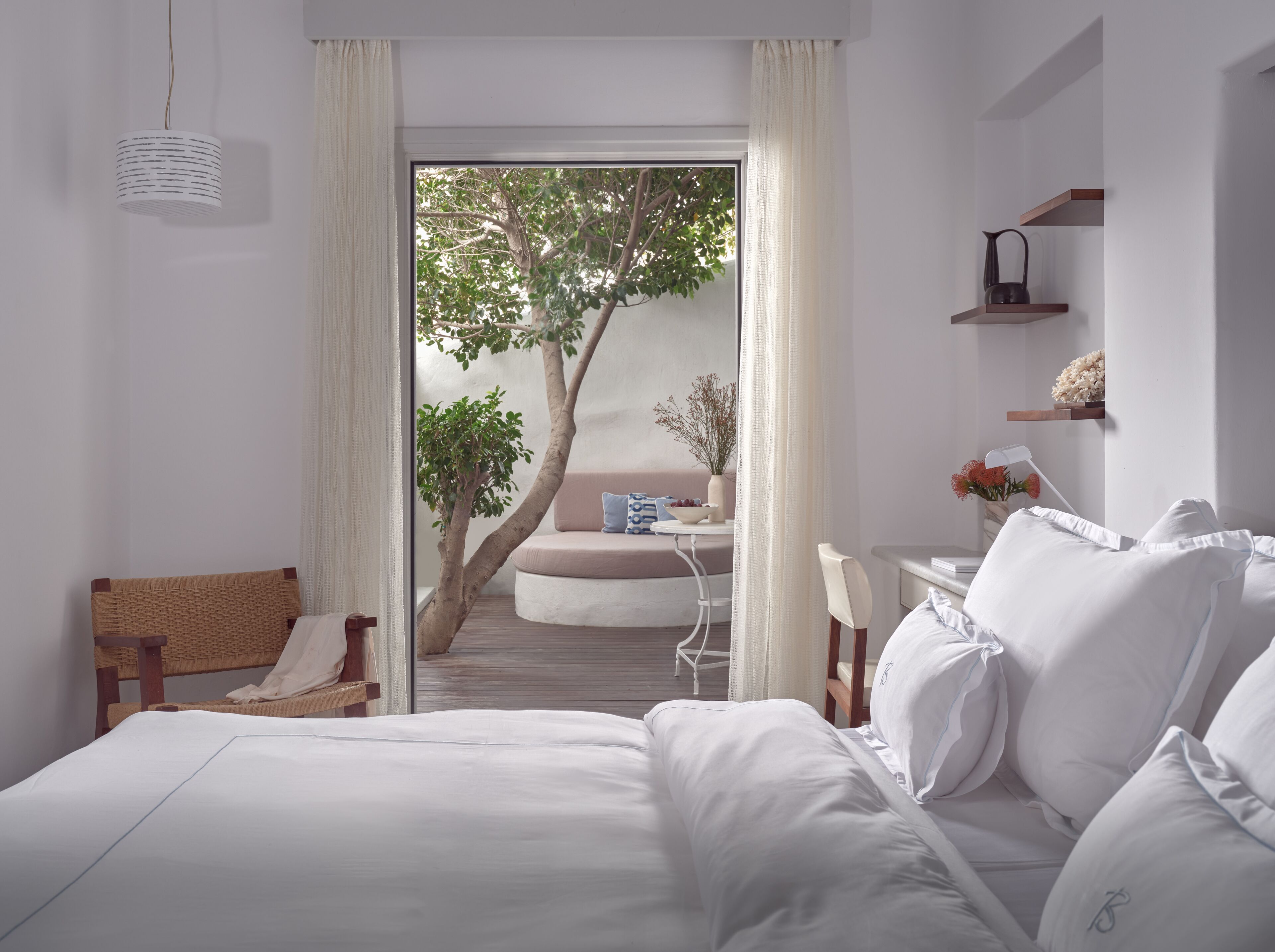 A bedroom with a bed, a wicker chair, a small table with a bowl, and a view of a balcony with a tree and a sofa.