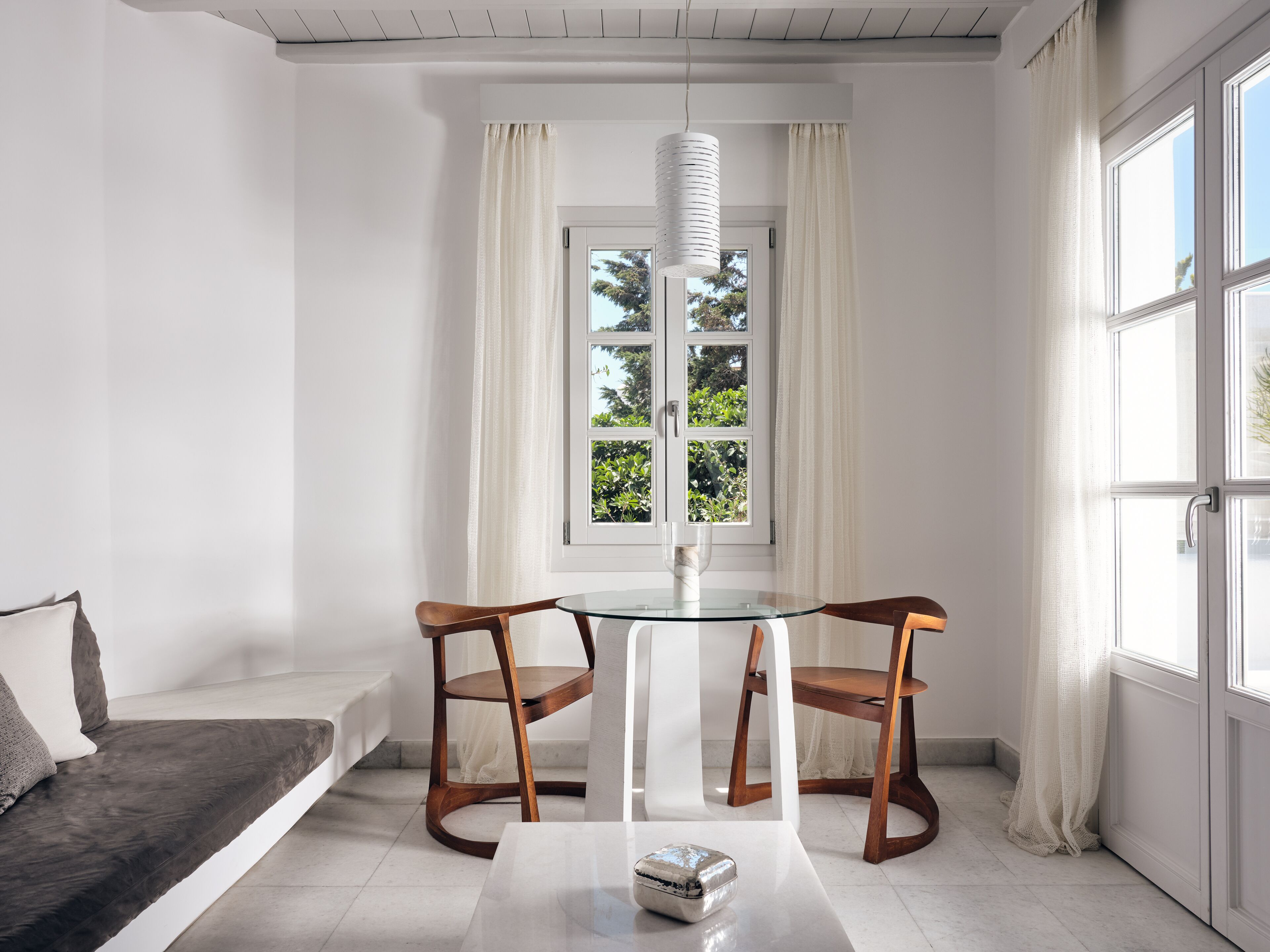 Una habitación moderna con una mesa de vidrio, dos sillas de madera, un sofá blanco y una ventana con cortinas.