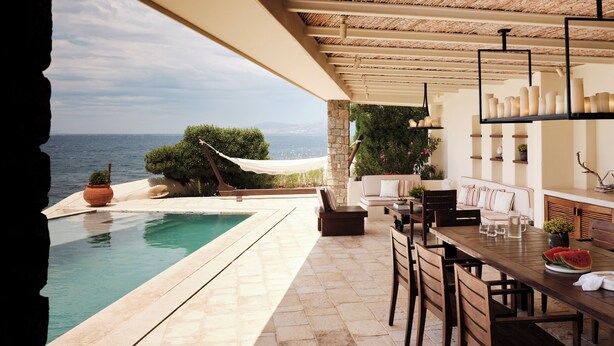 An outdoor pool area with a view of the sea, a wooden table with chairs, and a seating area with a sofa and cushions. - Belvedere Hotel - The Leading Hotels of the World  (Mykonos)